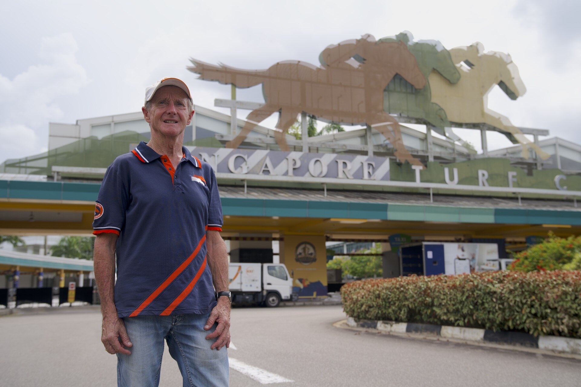An old man in a blue polo top stands near a large gate with three horse statues atop.