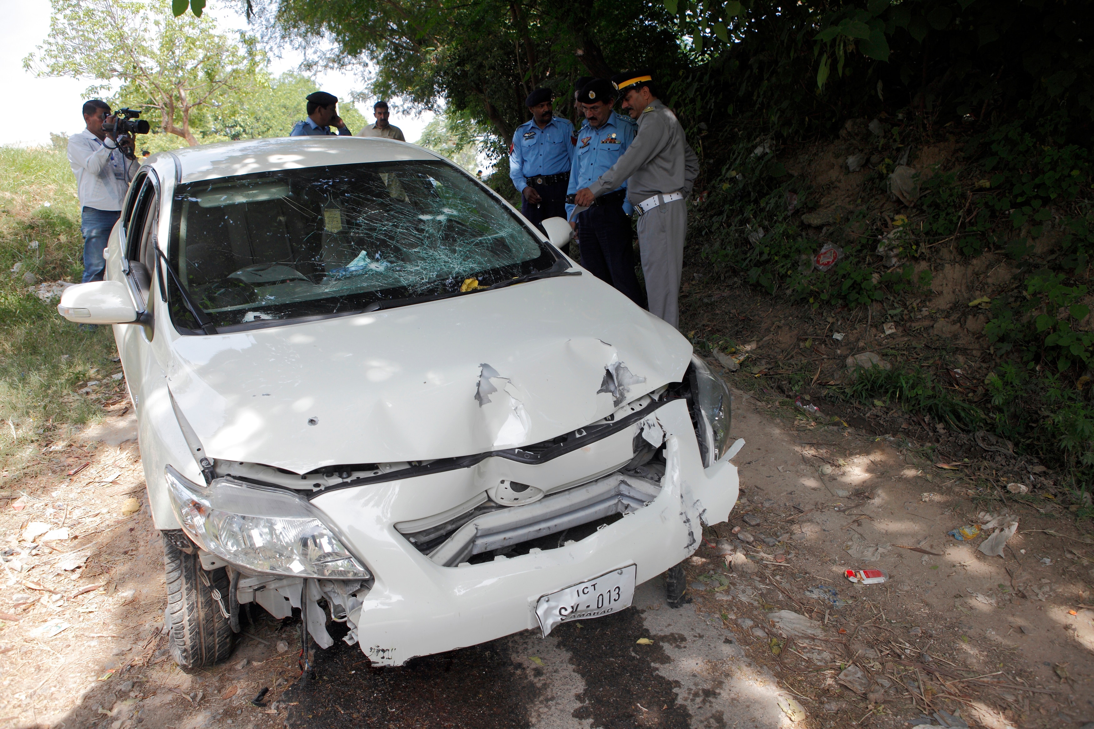 Agentes de segurança inspecionam um carro branco com o para-brisa quebrado e danificado em um ataque a tiros.