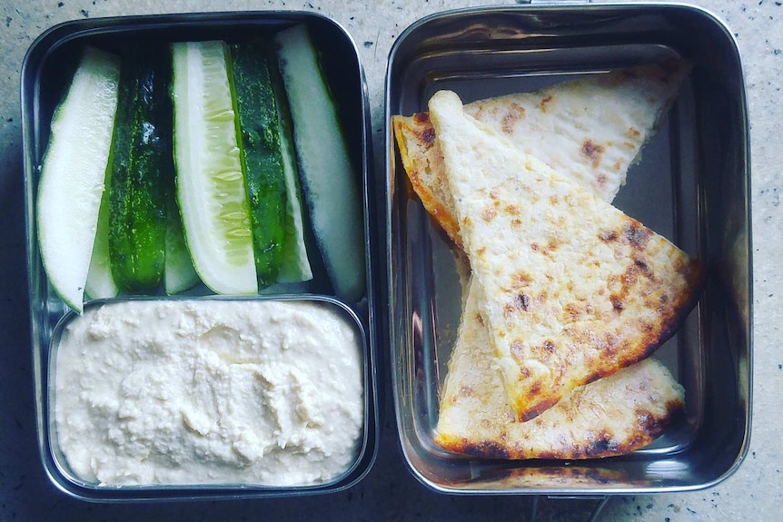 A metal lunch box with dips, cucumber and bread