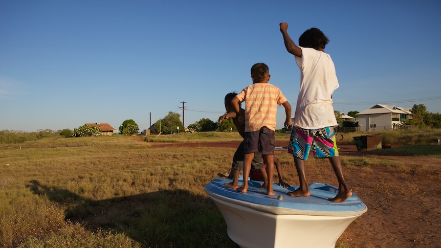 Three Aboriginal boys play on top of a boat parked on an area of grass, with houses and a road in the background.