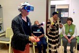 A lady wearing a virtual reality headset holding two game sticks and people standing around her cheering her on