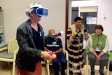 A lady wearing a virtual reality headset holding two game sticks and people standing around her cheering her on