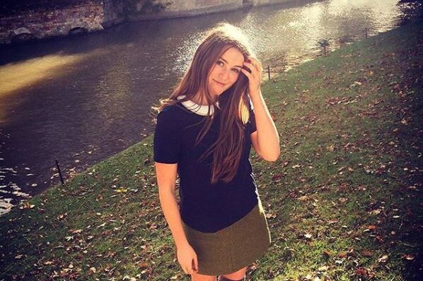 Caroline Calloway is seen standing on grass by a riverbank. She wears a khaki skirt and a black top with a white collar.