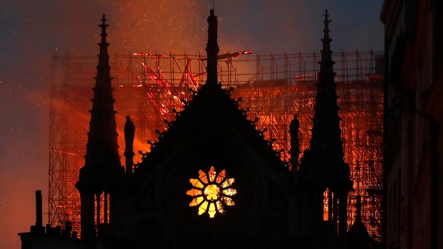 The silhouette of a cathedral is lit by embers and flames burning through a large gothic cathedral as the sky darkens