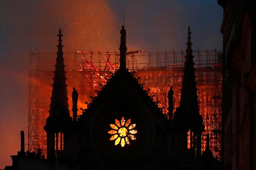 The silhouette of a cathedral is lit by embers and flames burning through a large gothic cathedral as the sky darkens