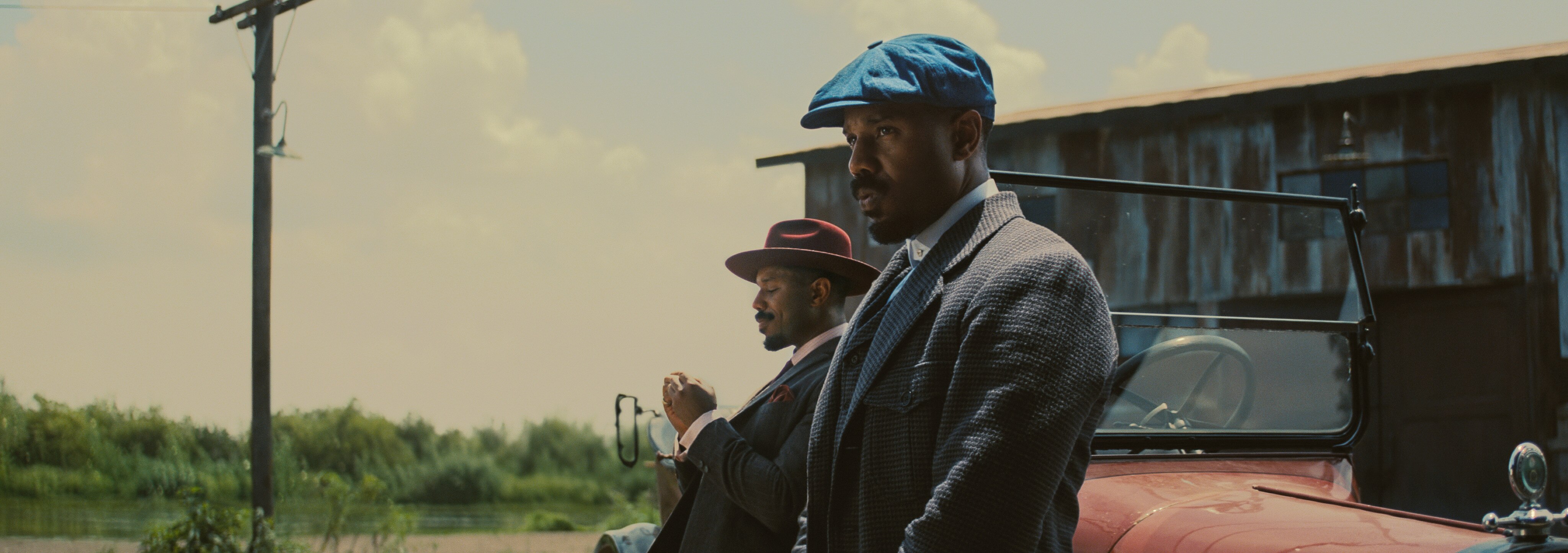 Two versions of Michael B Jordan in red and blue hats lean up against an old red topless ute