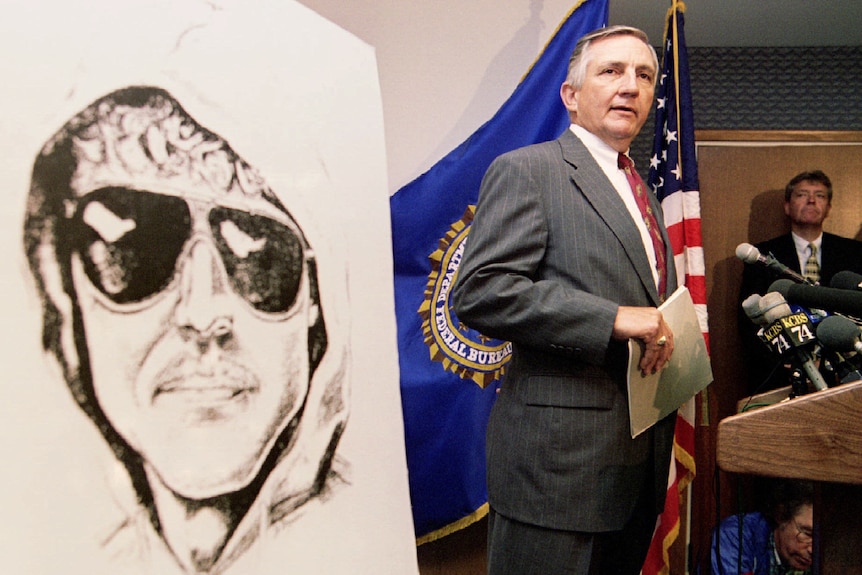 A man in suit and tie stands at a podium next to a large drawing of the Unbaomber, wearing sunglasses and hooded jumper