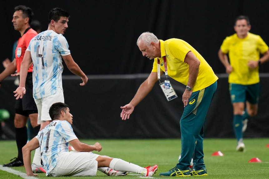Australia men's football coach Graham Arnold reaches down to help Argentina's Fernando Valenzuela off the ground.