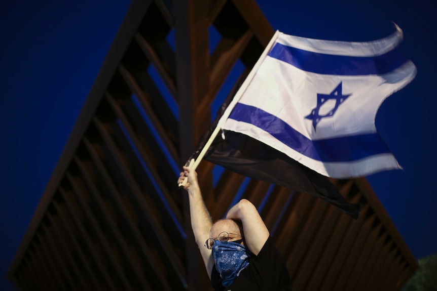 A person with their face covered waves an Israeli flag during a protest against Prime Minister Benjamin Netanyahu.