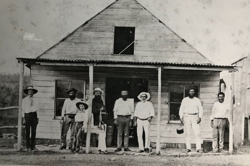 A black and white grainy picture of several people standing in front of a small wooden building