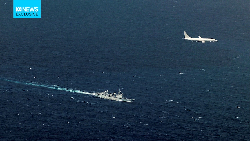 An aerial photo showing a Navy ship in the open ocean, with a military jet flying overhead.