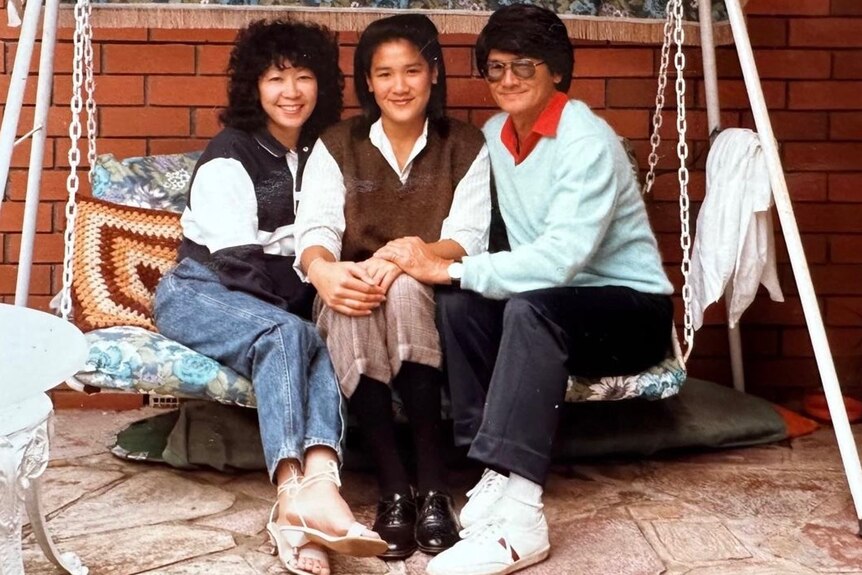 Kylie Kwong as a teenager sits on an outdoor swing chair next to her mum and dad 