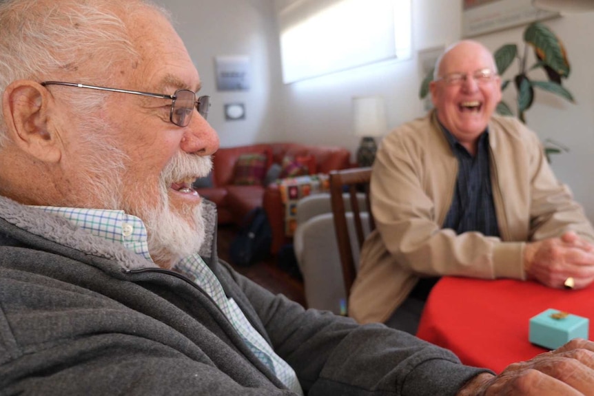 Two older men sit at a kitchen table smiling and laughing.
