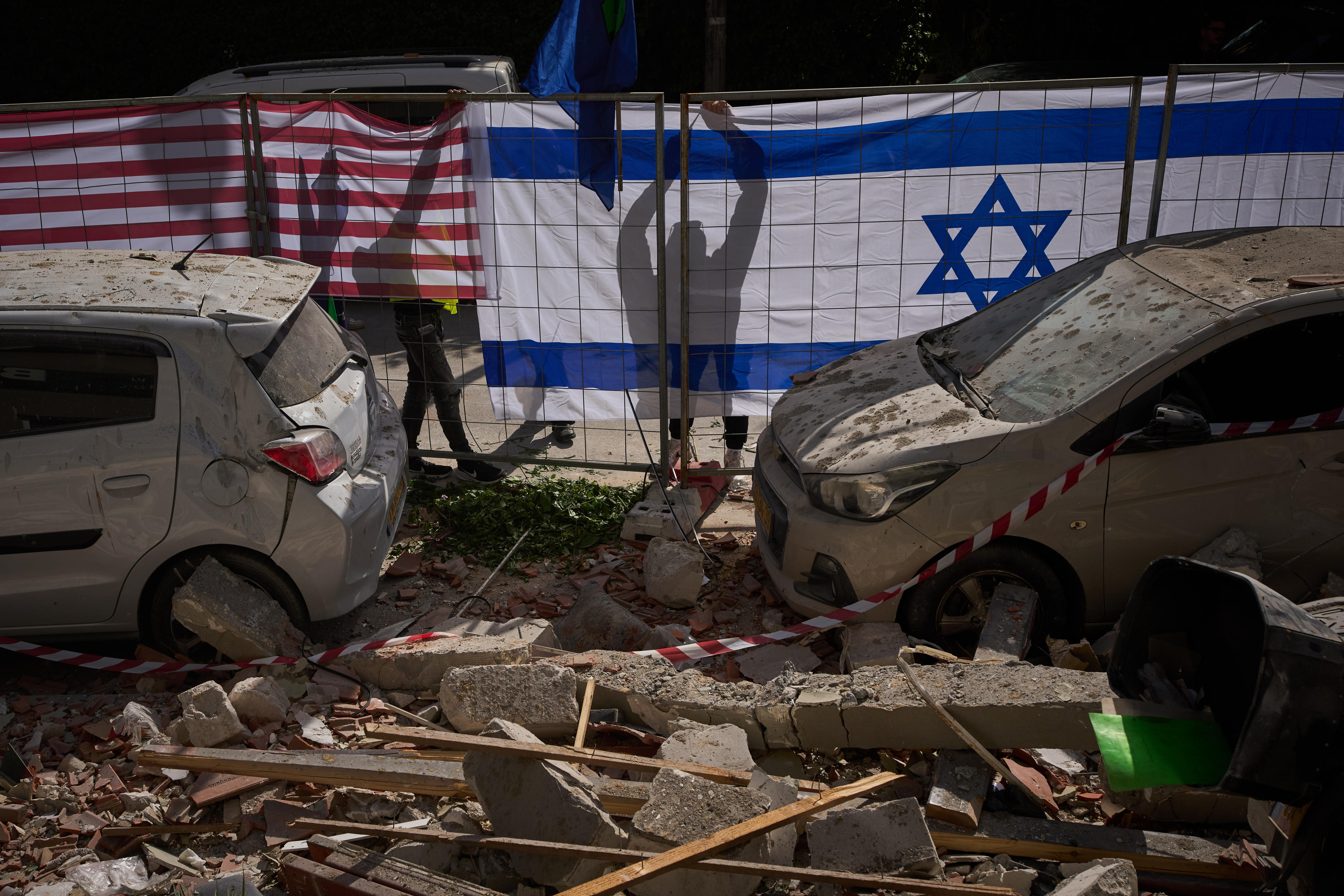 Israeli and American flags draped on hoardings behind two damaged cars from an Iranian missile strike.