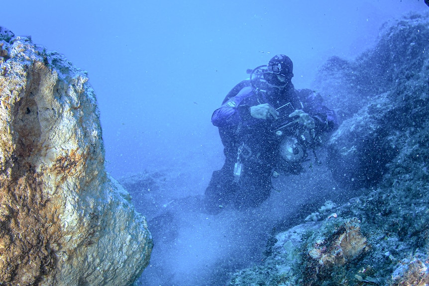 A diver underwater swims past coral and rocks.