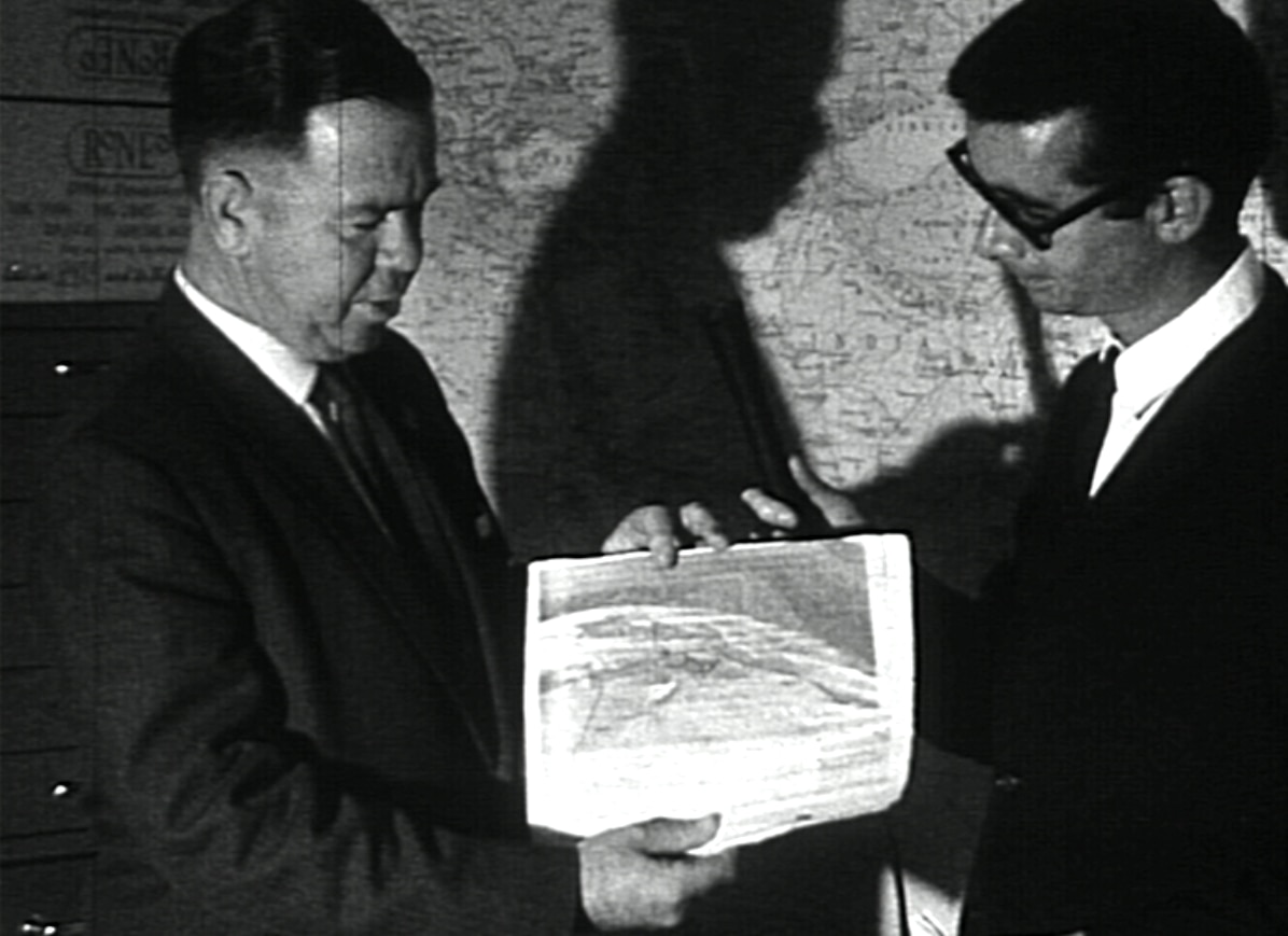 Um meteorologista e um jornalista observam uma foto granulada de um satélite, impressa em papel.