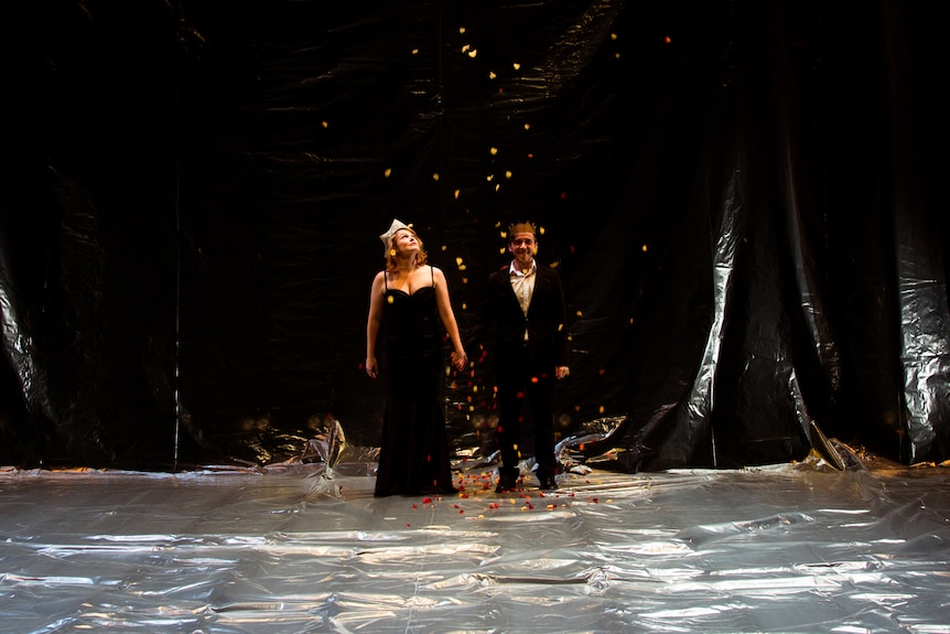 A blonde woman in her early 30s holds hands with a man in his 30s, each formally dressed, as confetti falls from the sky
