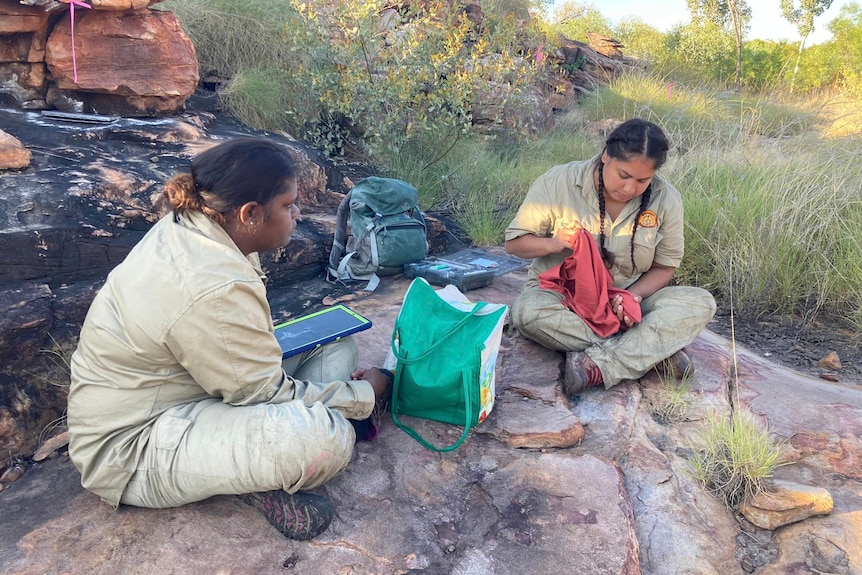 Two women in khaki sit on a rock with a shopping bag, backpack and a sack with a bandicoot inside.