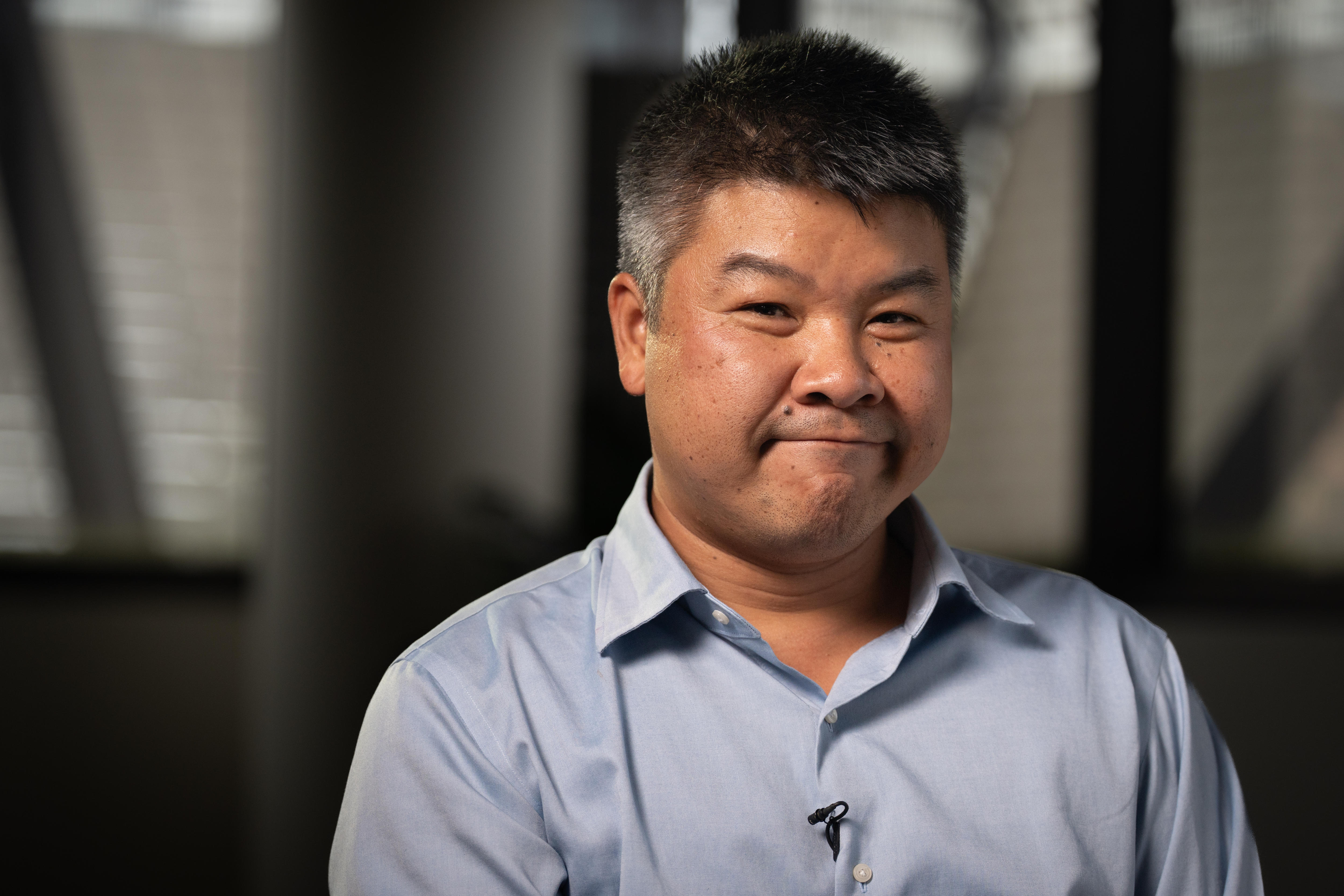 Um homem de Hong Kong com sorriso fechado, cabelos pretos/grisalhos, vestindo uma camisa de botão azul claro e um escritório escuro atrás dele.
