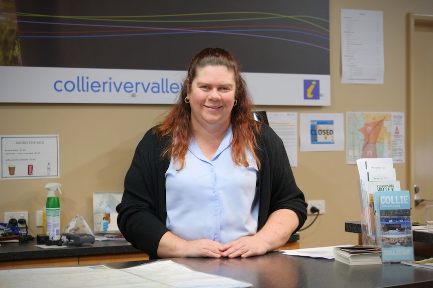 A woman wearing a blue shirt and black jumper stands behind a desk