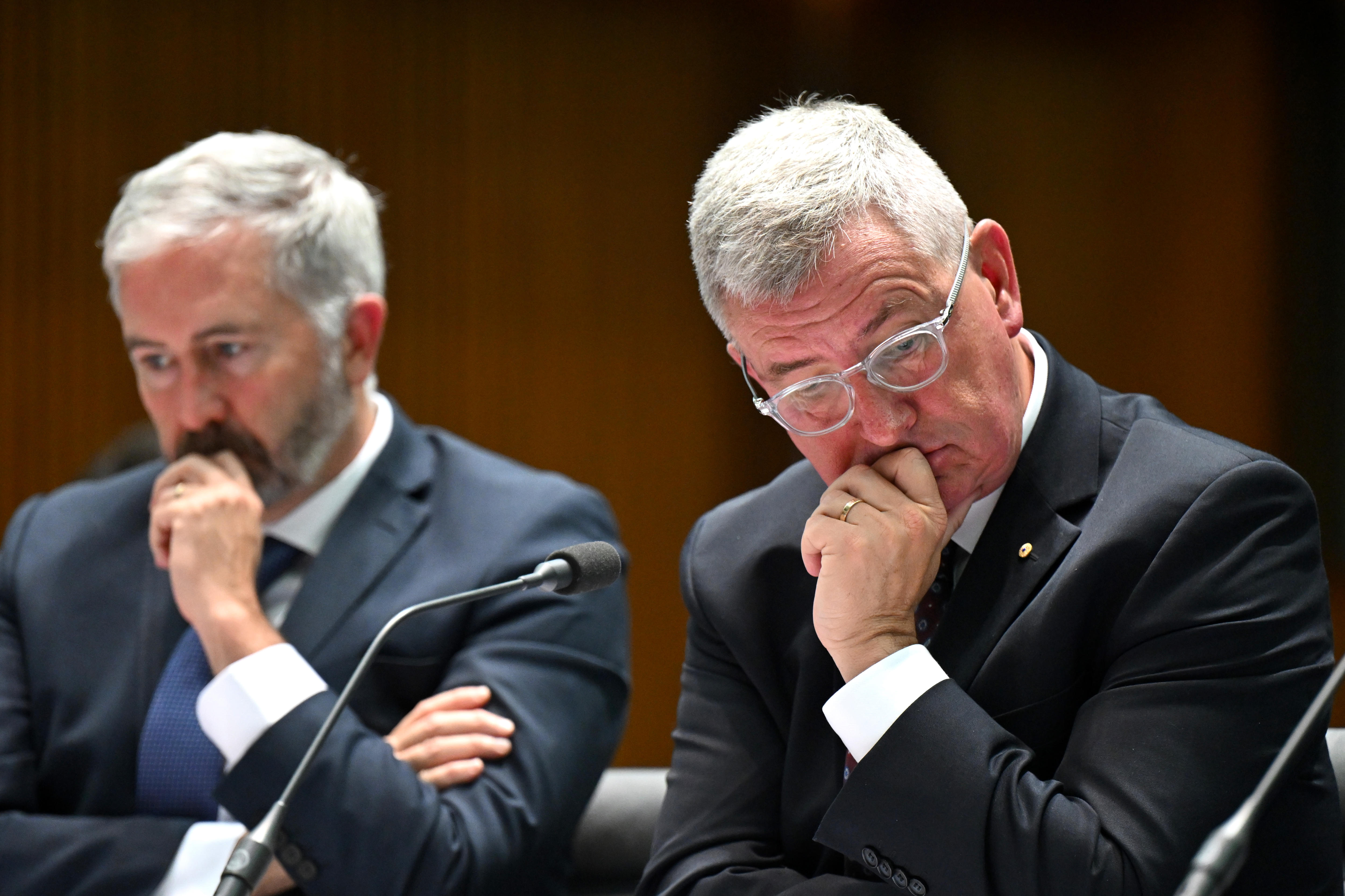 Dois homens brancos de meia-idade, cabelos brancos e ternos azul-escuros estão sentados e parecendo preocupados.