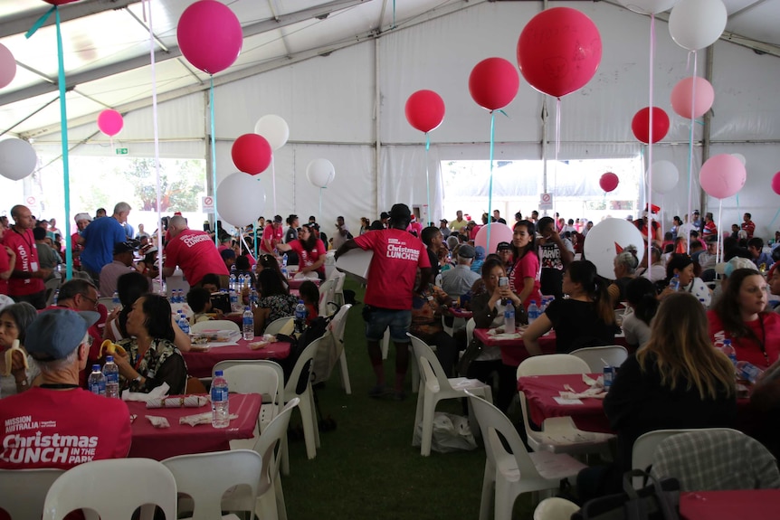 People sit on white plastic chairs at red tables decorated with balloons inside a large marquee