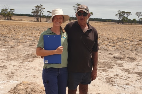 Freya Spencer stands left next to a farmer in an open field. She has a hat on and holds a clipboard. The field is brown.