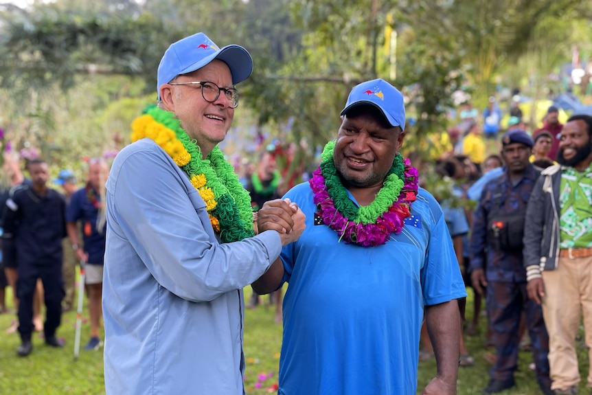 Albanese and Marape, wearing leis, clasp hands together and smile, with a crowd behind them.