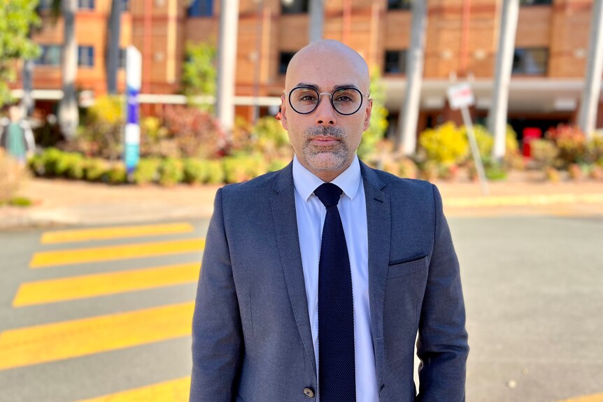 A bald man standing in front of a brick hospital. He is wearing a suit and dark framed glasses.