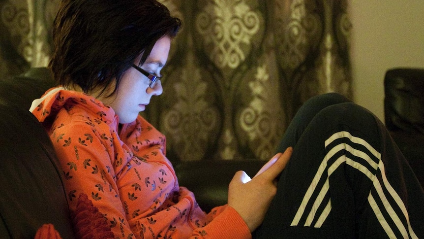 A girl in a red jumper and black tracksuit pants sits on a couch indoors at night looking at her mobile phone.