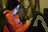 A girl in a red jumper and black tracksuit pants sits on a couch indoors at night looking at her mobile phone.