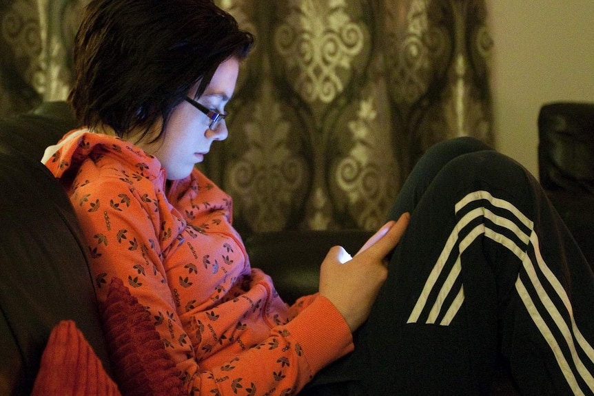 A girl in a red jumper and black tracksuit pants sits on a couch indoors at night looking at her mobile phone.