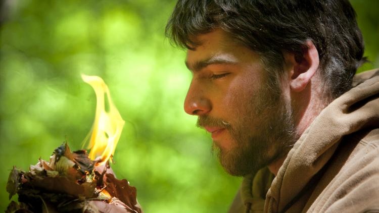 A man holds a tinder bundle that has caught alight near his face.