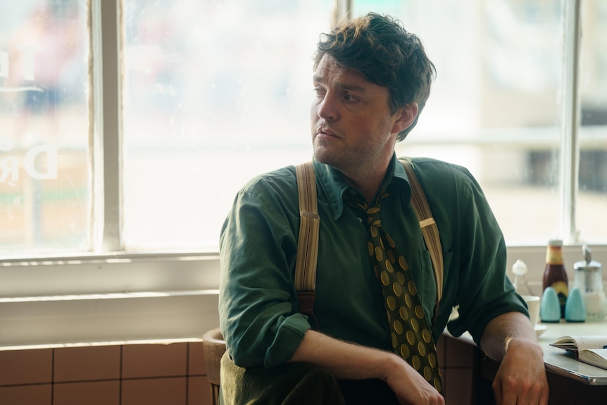 A man in his early 40s in a 50s-style shirt and suspenders, sits in a cafe, looking wistfully to the side