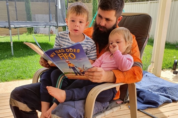 A man in a high-vis vest reads a book called 'My Dad does FIFO' to two small kids on his lap in a chair in a backyard.