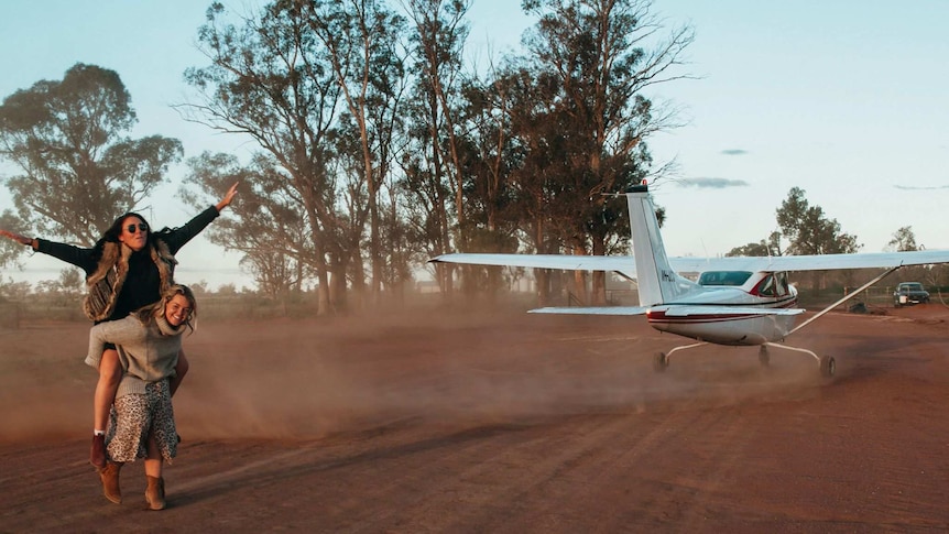 women riding piggy back on a dusty runway in front of a plane.