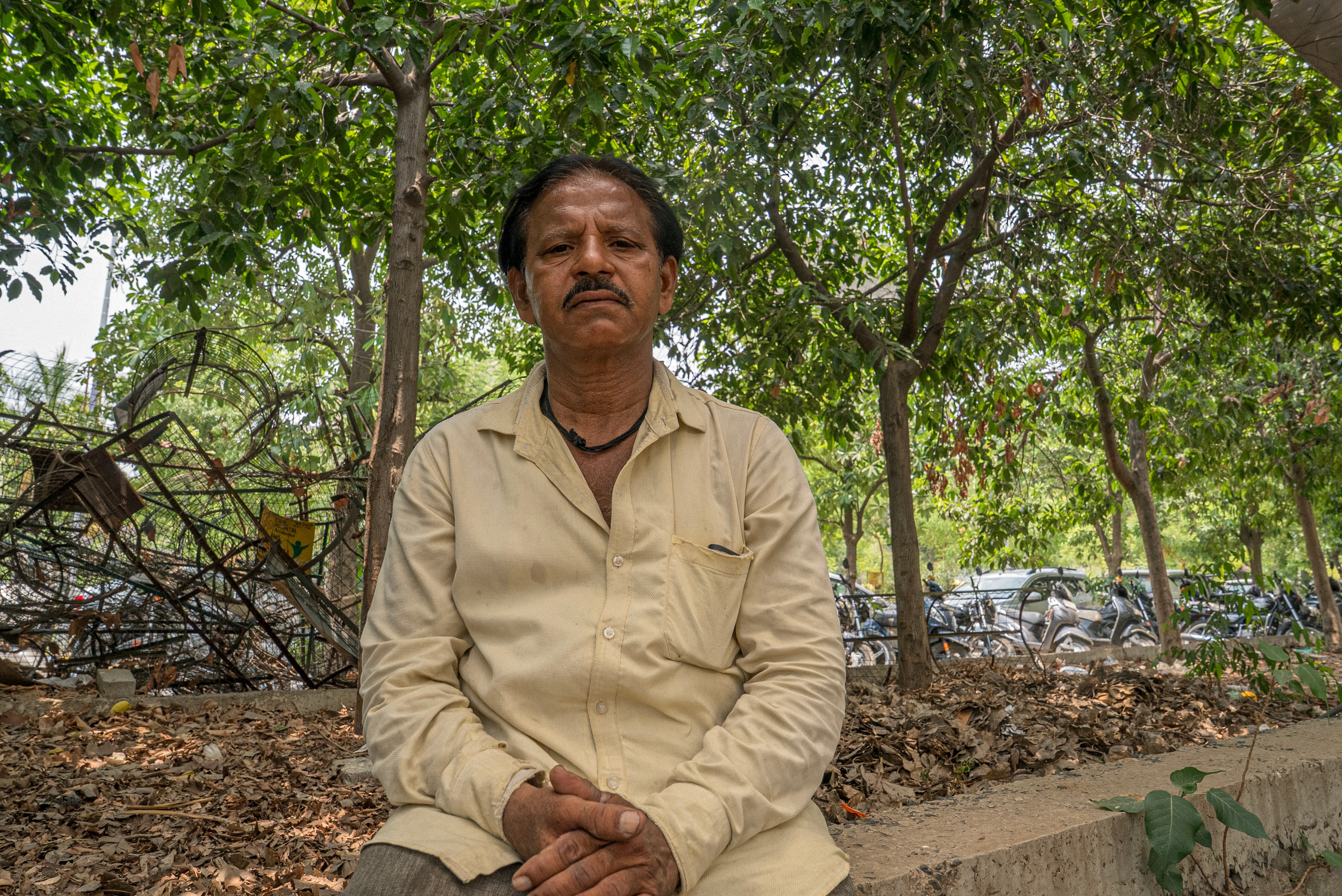 Indian man wearing a yellow shirt sitting on a concrete wall with trees in the background.