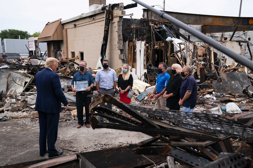 A man faces a group of people wearing masks who are standing in front of a badly damaged building.