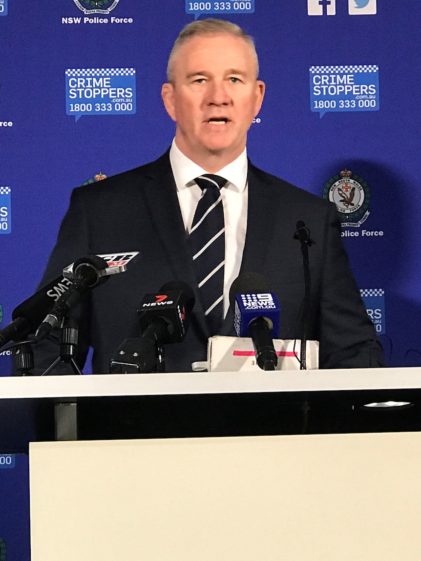 A man wearing a suit stands before microphones in front of a wall of NSW Police logos.