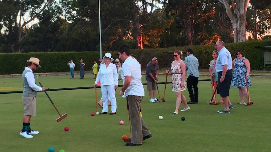 People play croquet in colourful outfits, very relaxed feel on greens with tall trees in background.