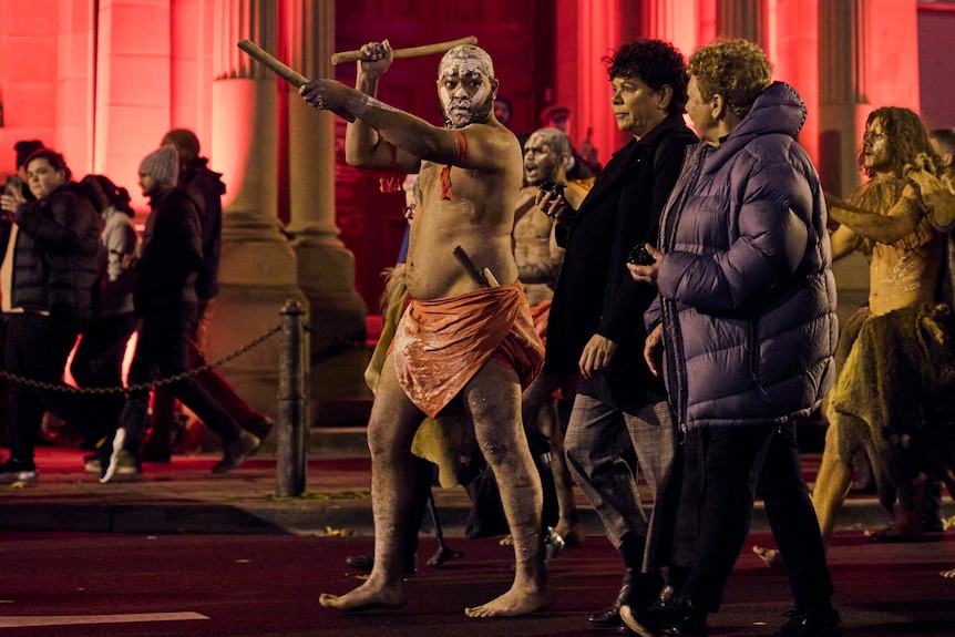 A group of First Nations performers carrying traditional objects walk among a crowd of people wearing warm clothes on a street.