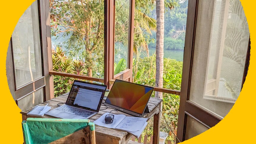 A laptop sits open on a desk before an open window looking at over a river.