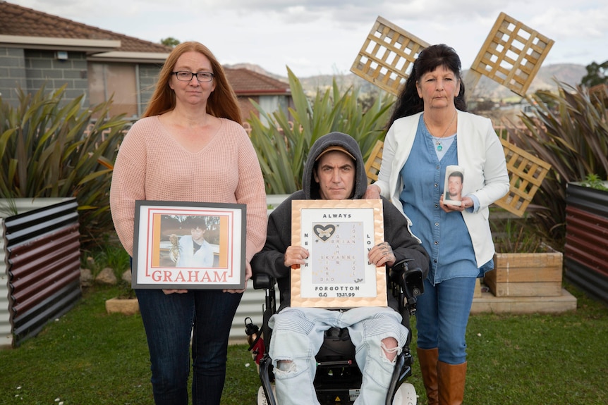 A woman with red hair stands next to a seated man and another woman holding photo frames