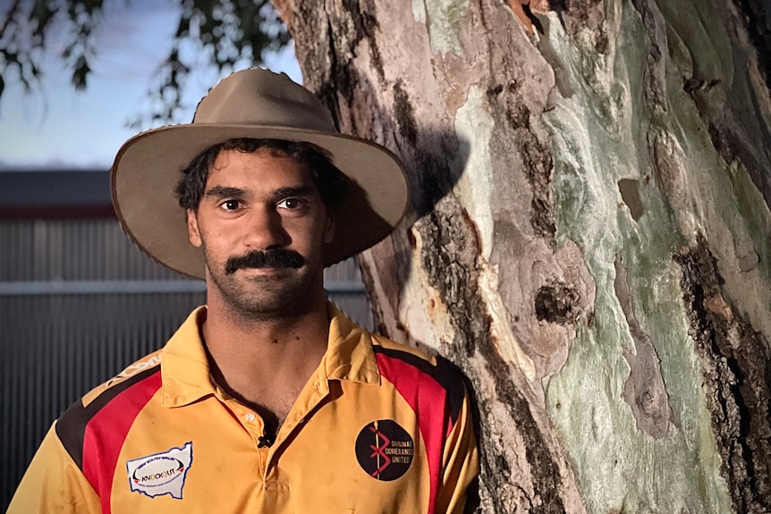 Chris McHughes stands next to a tree, he wears a wide-brimmed hat and a yellow, black and red shirt.