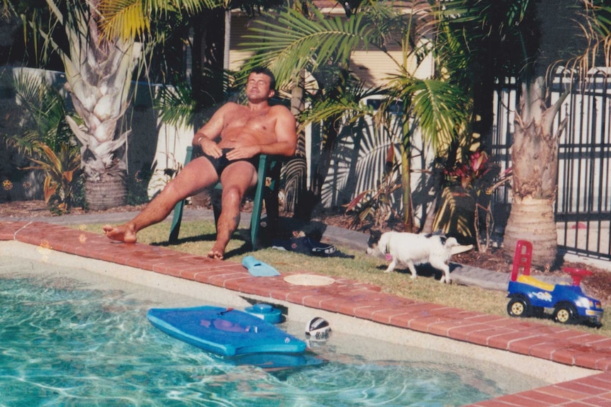 A man sits in a plastic outdoor chair and suns himself next to a swimming pool.
