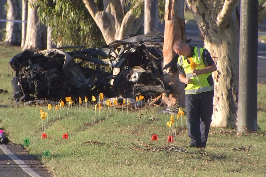A mangled crashed car next to a tree, while a police officer wearing high vis puts down yellow flags.