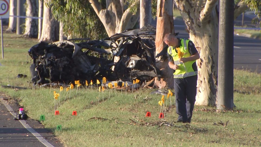 A mangled crashed car next to a tree, while a police officer wearing high vis puts down yellow flags.