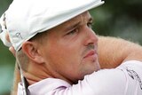 Bryson DeChambeau holds a pose with his driver wearing a light coloured shirt and a white hat