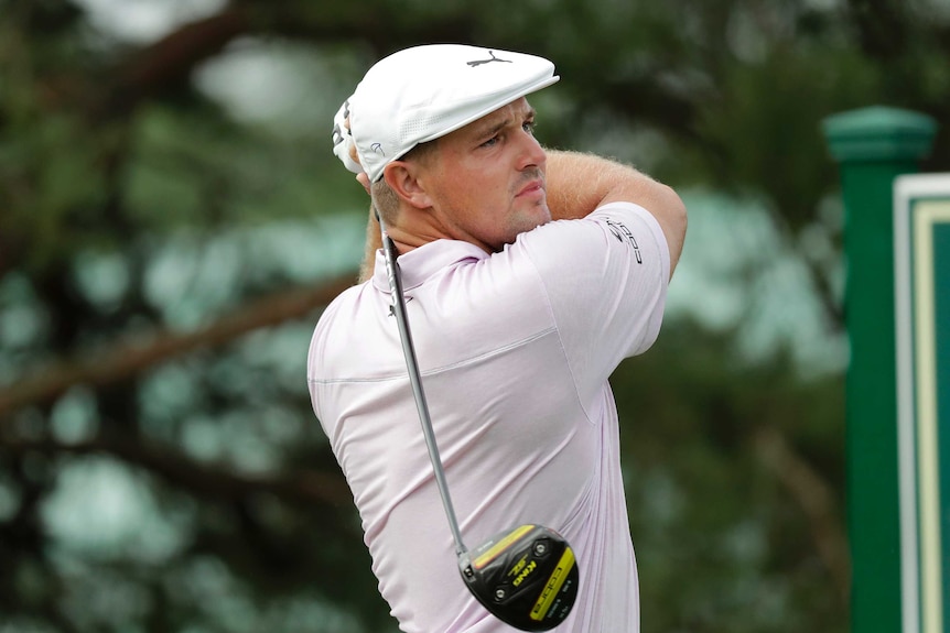 Bryson DeChambeau holds a pose with his driver wearing a light coloured shirt and a white hat