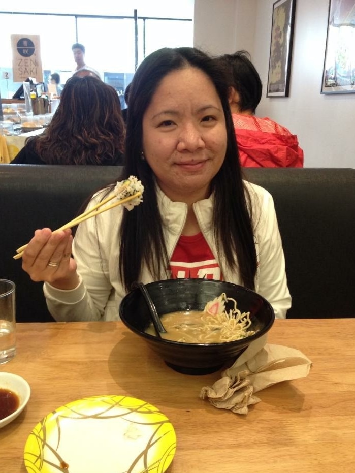 A women sits in a restaurant booth and smiles with a bowl of noodles while holding sushi with chopsticks.
